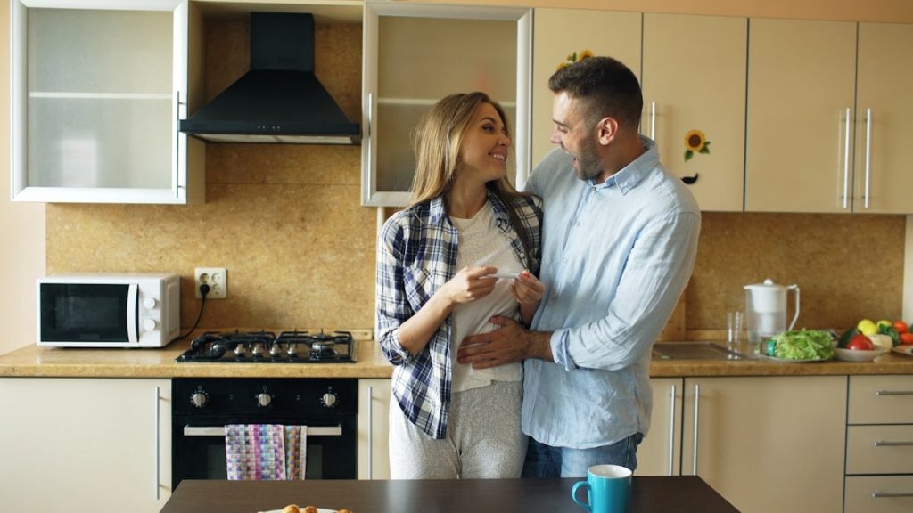 Attractive girl shows pregnancy test result to her boyfriend and surpise him at the kitchen . Loving couple embrace and kissing at home because of good news