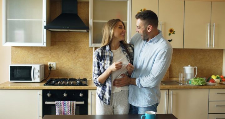 Attractive girl shows pregnancy test result to her boyfriend and surpise him at the kitchen . Loving couple embrace and kissing at home because of good news